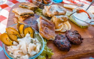 Plato nacional albanés con carne a la parrilla, pastas crujientes y verduras frescas, servido en bandeja de madera.