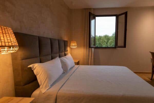 Acogedora habitación de hotel con cómoda cama, lámpara de madera y vistas a la naturaleza en Albania. Perfecta para unas vacaciones relajantes en la aventura albanesa.