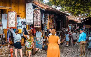 Colorida artesanía albanesa en un mercado tradicional de Albania, con tejidos, ropa y recuerdos locales, rodeado de edificios históricos de madera y un ambiente animado.