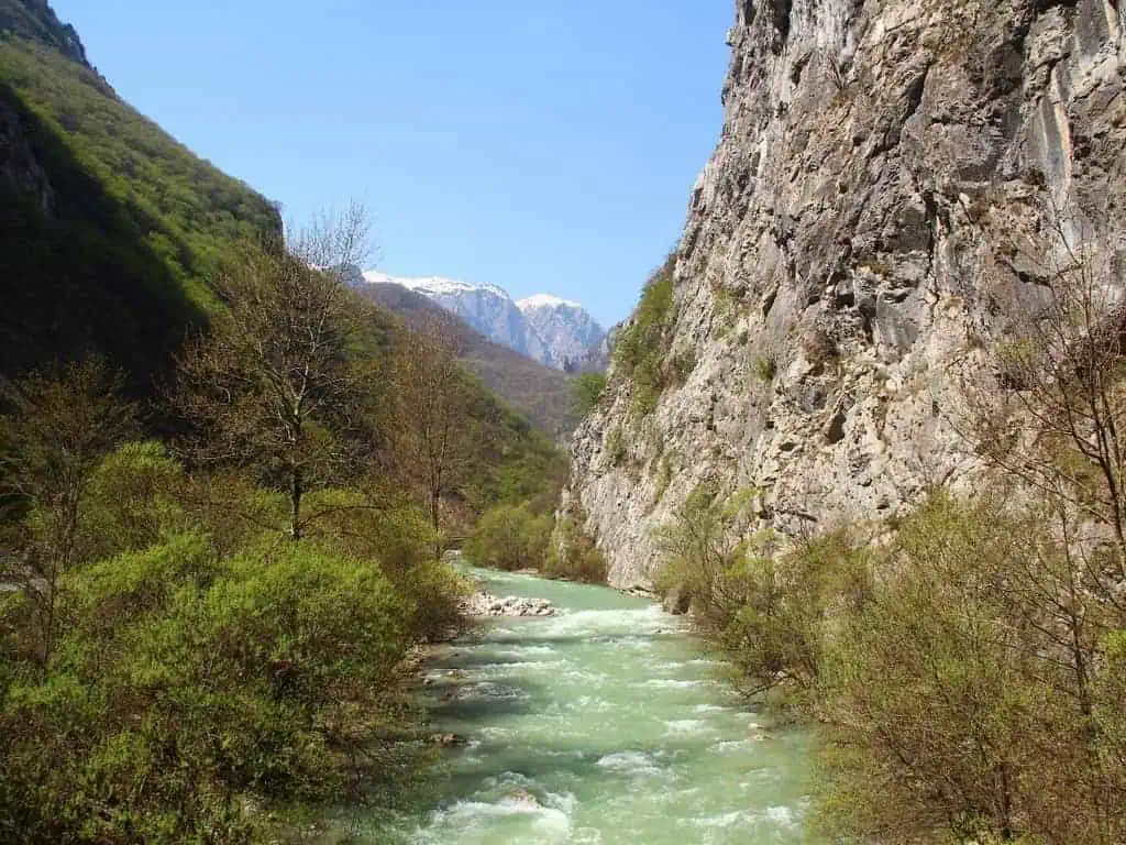 Landelijk rivierlandschap in Albanië met groen water, omringd door hoge bergen en groene dennenbomen, onder een helderblauwe hemel, ideaal voor natuurliefhebbers en avontuurlijke vakanties.