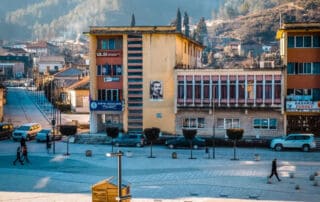 Aventura Albania, paisaje urbano con edificios modernos, arquitectura socialista y paisaje montañoso de fondo. Ambiente urbano animado con peatones y coches, escenas típicas de Albania.