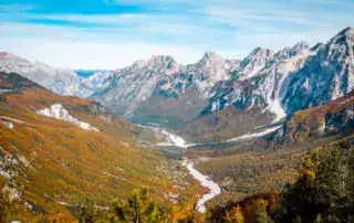 Impresionante paisaje montañoso albanés con picos nevados, densa vegetación y un río que fluye por el valle. Perfecto para los amantes de la naturaleza y el senderismo.