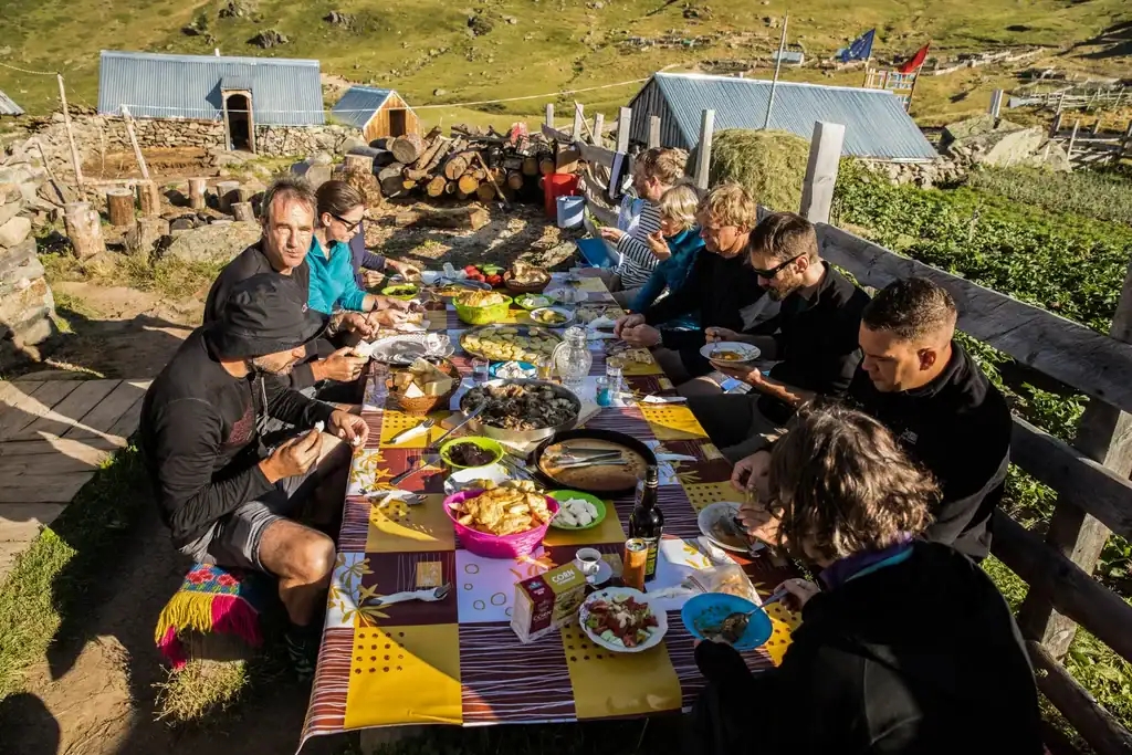 Adventure Albania, authentic food in picturesque nature, muestra a un grupo de personas disfrutando de un picnic tradicional albanés al aire libre, rodeados de montañas y arquitectura histórica.