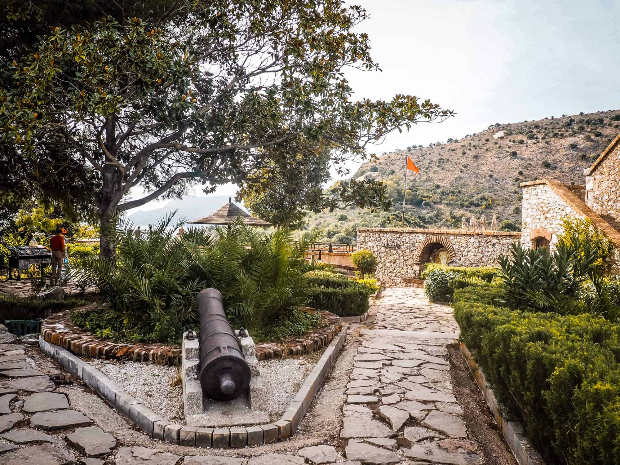Historischer Ort in Albanien mit einer alten Kanone, umgeben von grünen Pflanzen und malerischen Steingebäuden, bei sonnigem Wetter und Blick auf die Berge.