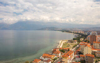 Fotografía de alta calidad de Albania, que muestra la costa con encantadoras casas e impresionantes montañas al fondo, ideal para auténticos viajes por los Balcanes y turismo de naturaleza.