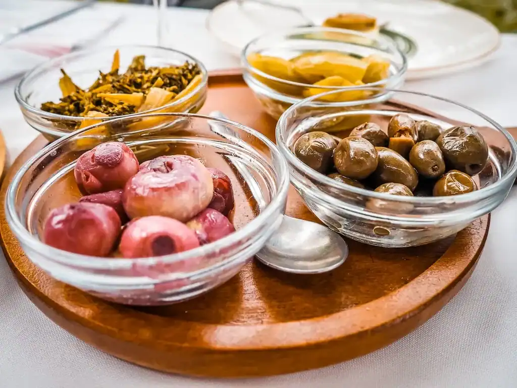 Coloridas tapas albanesas con verduras encurtidas y aceitunas en tarros sobre una tabla de madera, típicas de la cocina regional.