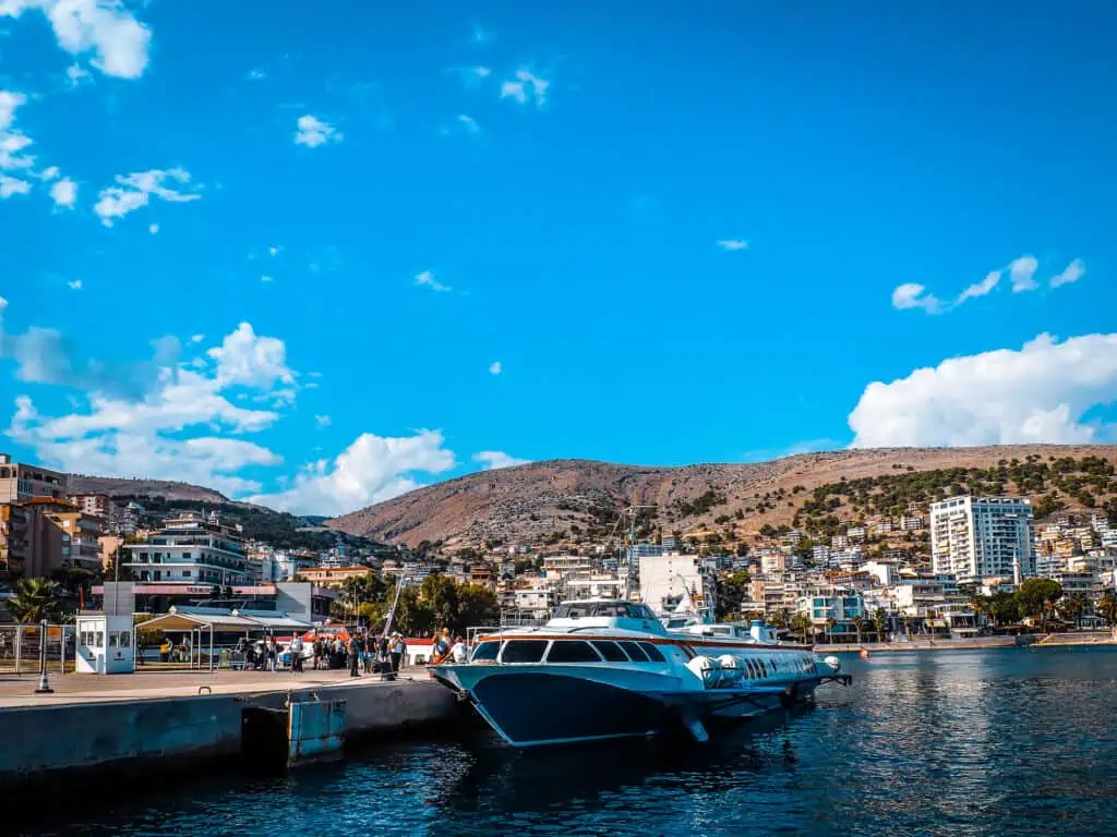 Beeleuchtete Hafenpromenade in Albanien, mit Yachten, Stadtgebäuden und Bergen im Hintergrund, sonniges Wetter und klare blaue Himmel.