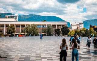 Vista frontal de la plaza central de Tirana, Albania, con edificios modernos, montañas al fondo y gente explorando la ciudad, ideal para fotos de viajes e información turística.