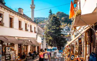 Coloridas calles del casco antiguo de Albania, con tiendas locales, turistas y pintorescos paisajes de montaña, ideales para vivir aventuras y experiencias culturales en Albania.