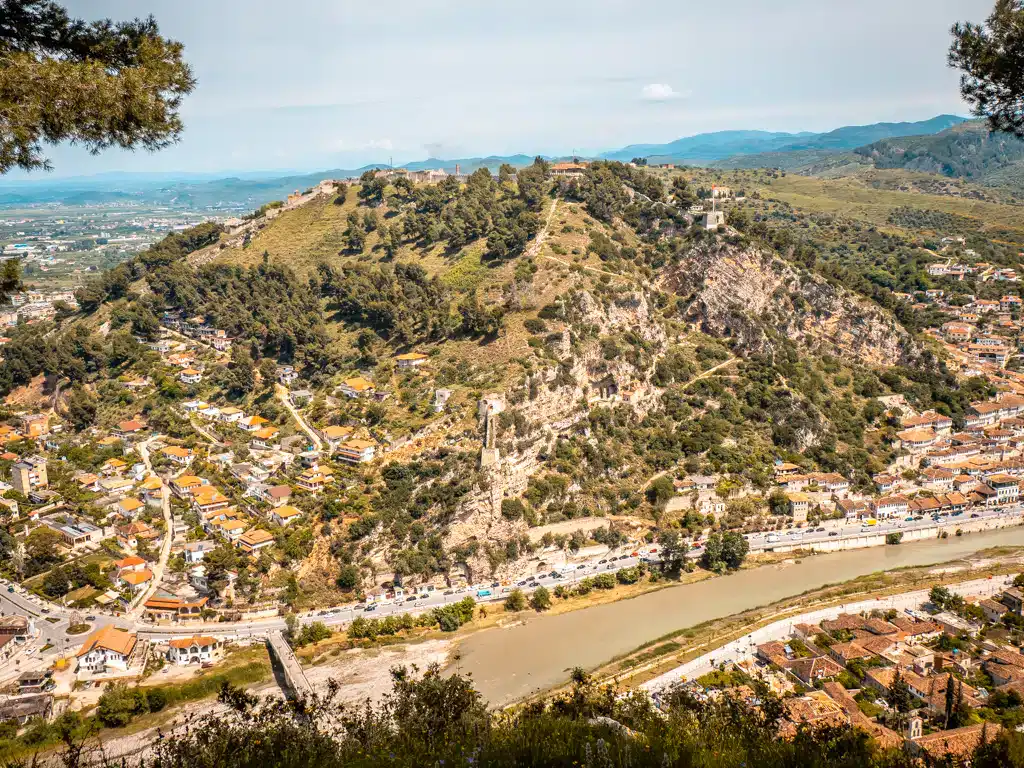 Aventura Albania: Pintoresca vista de la ciudad bajo la colina con castillo, rodeada de montañas, río y arquitectura tradicional, perfecta para los amantes de los viajes de naturaleza y cultura.